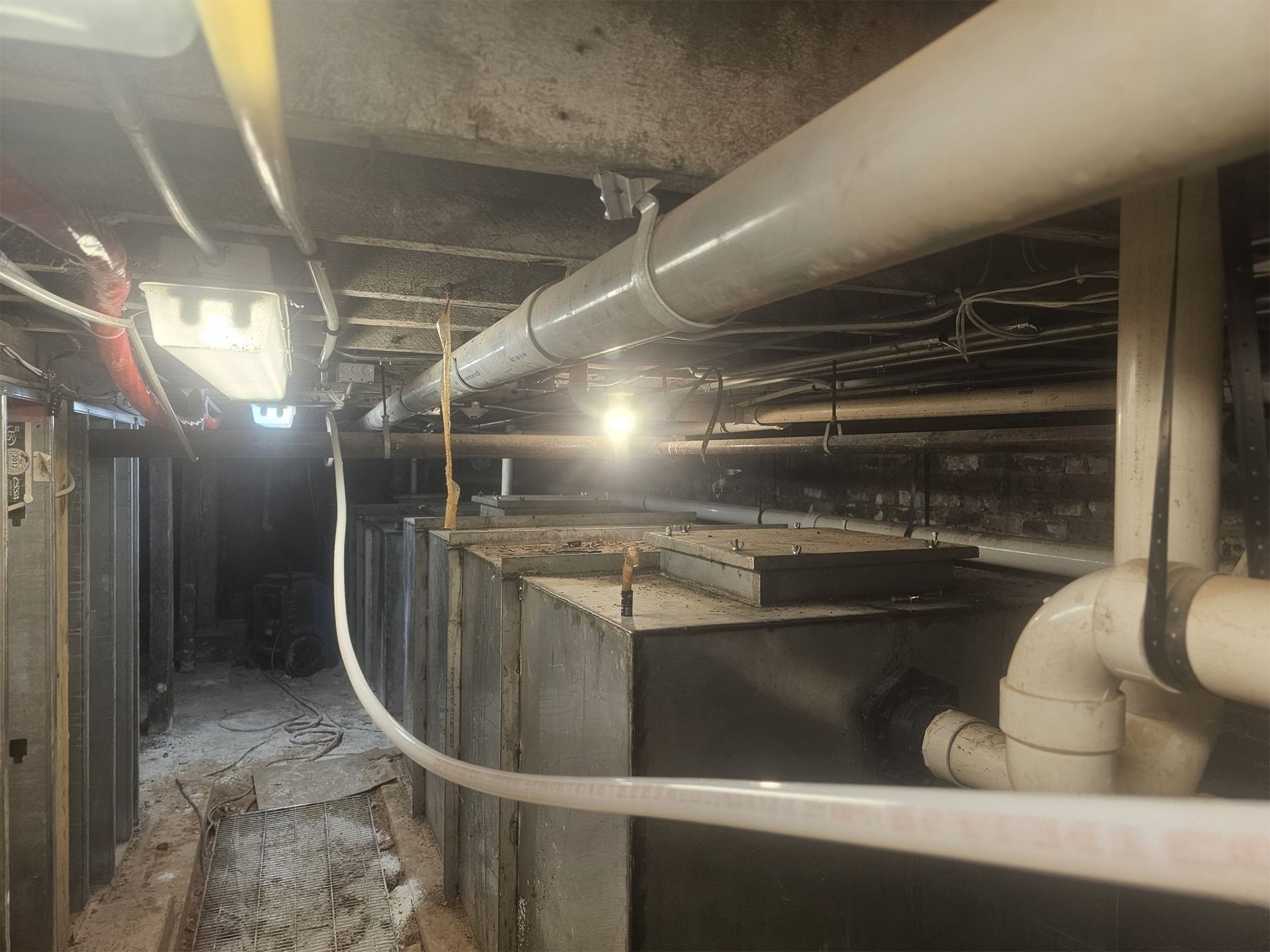 Dim basement utility space with exposed pipes, ducts, and concrete walls lit by bright overhead lights