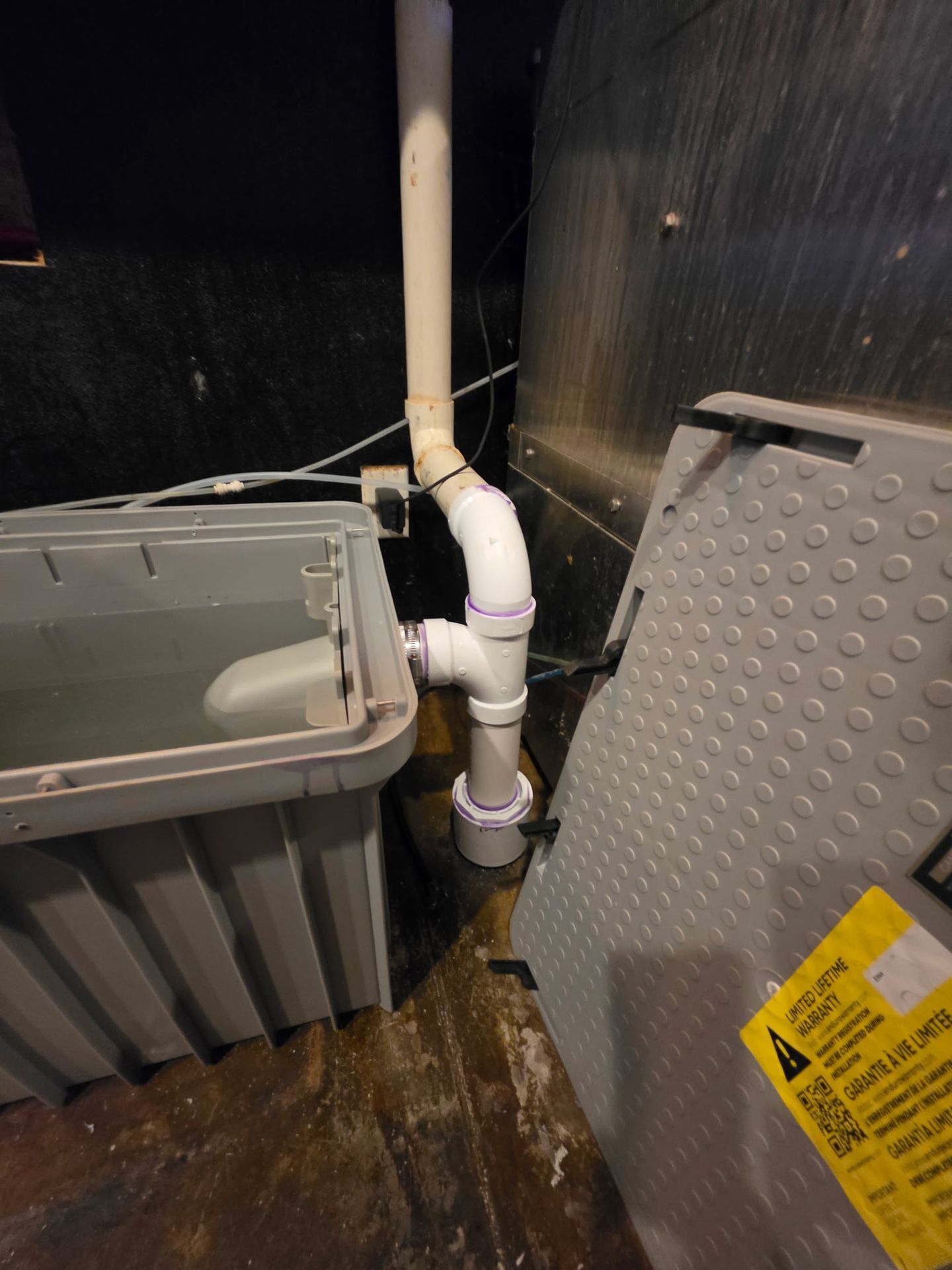 White plumbing pipes beside a gray bin and a silver panel in a cramped utility corner