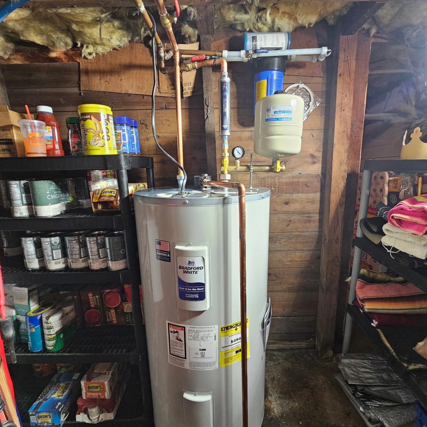 Utility room with a white water heater, shelves of supplies, and exposed pipes in a basement-like space