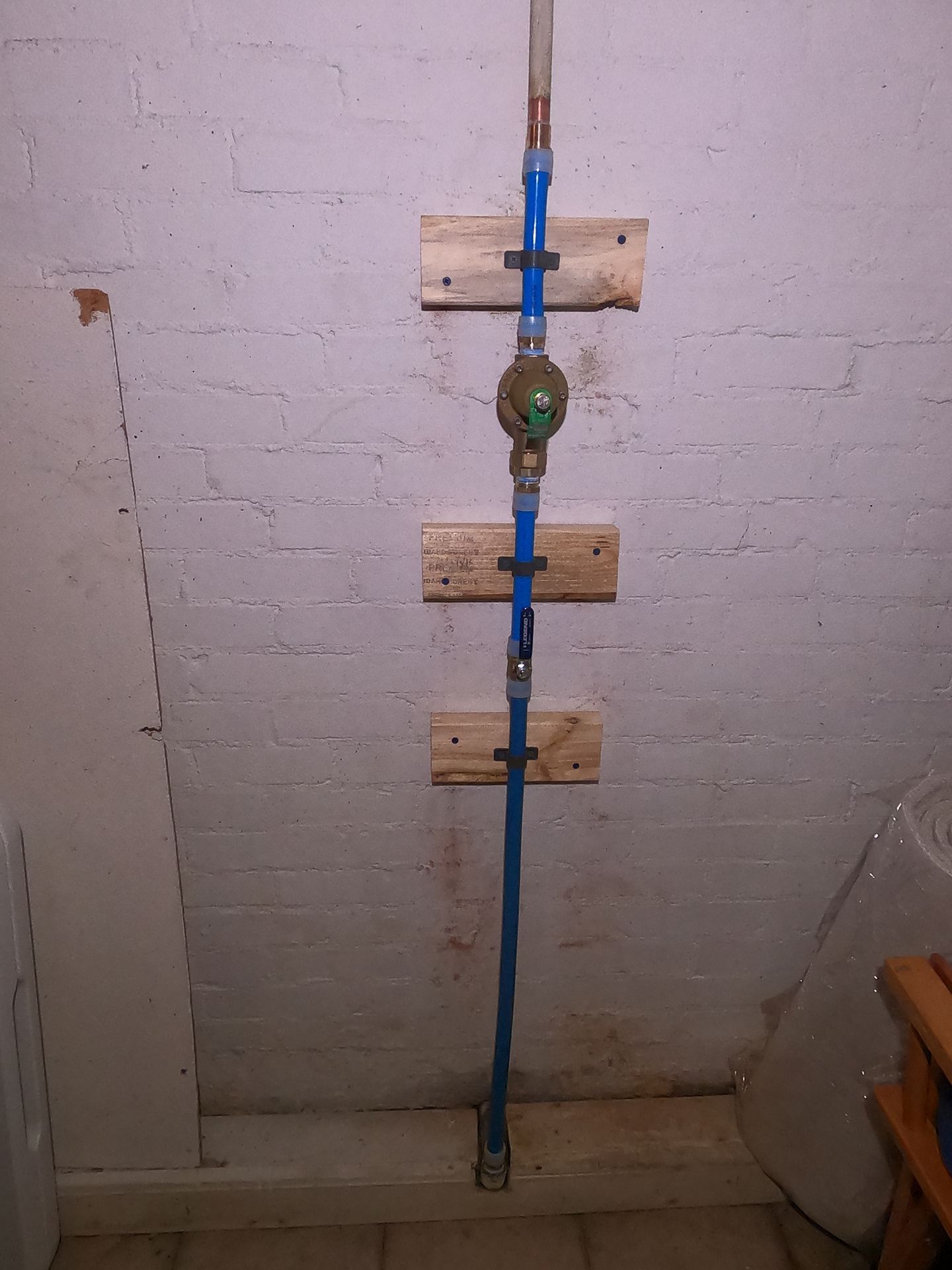 Blue vertical pipe with a round valve mounted on a white brick wall, secured by wooden brackets.