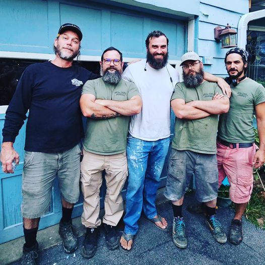 Five men standing together outdoors in front of a blue building, smiling with arms around each other.