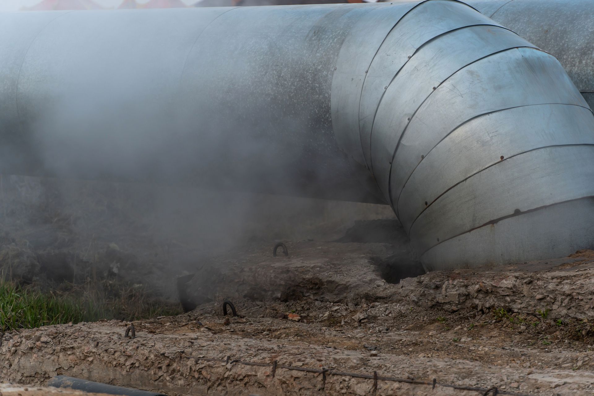 Large silver industrial pipe venting steam over muddy ground in an outdoor worksite