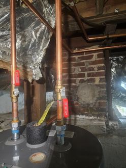 Copper pipes and valves above a black water heater in a basement with brick walls.