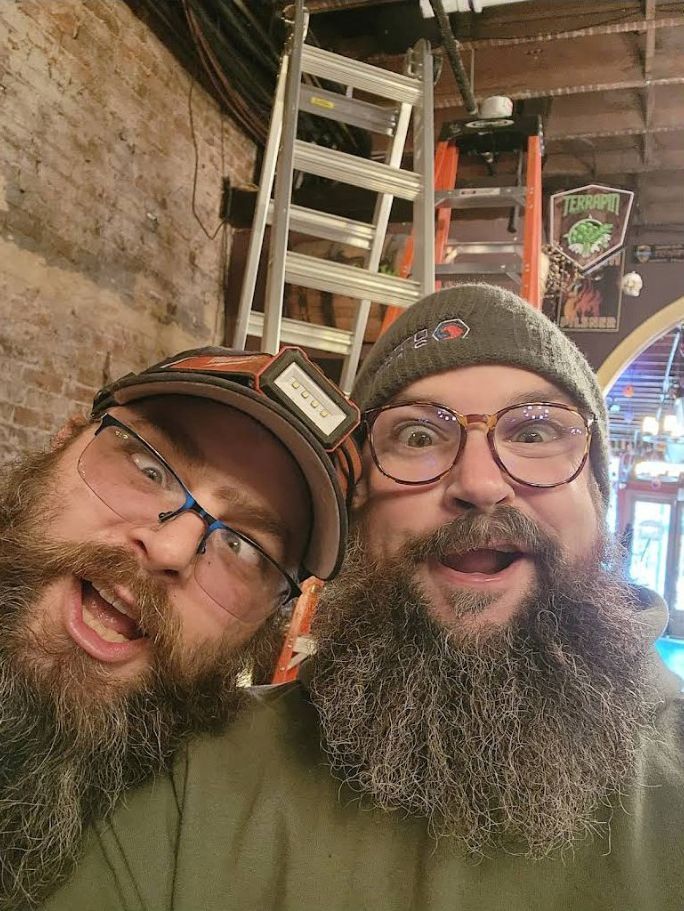 Two bearded men smiling in a rustic bar, one in a cap and one in a beanie and glasses.