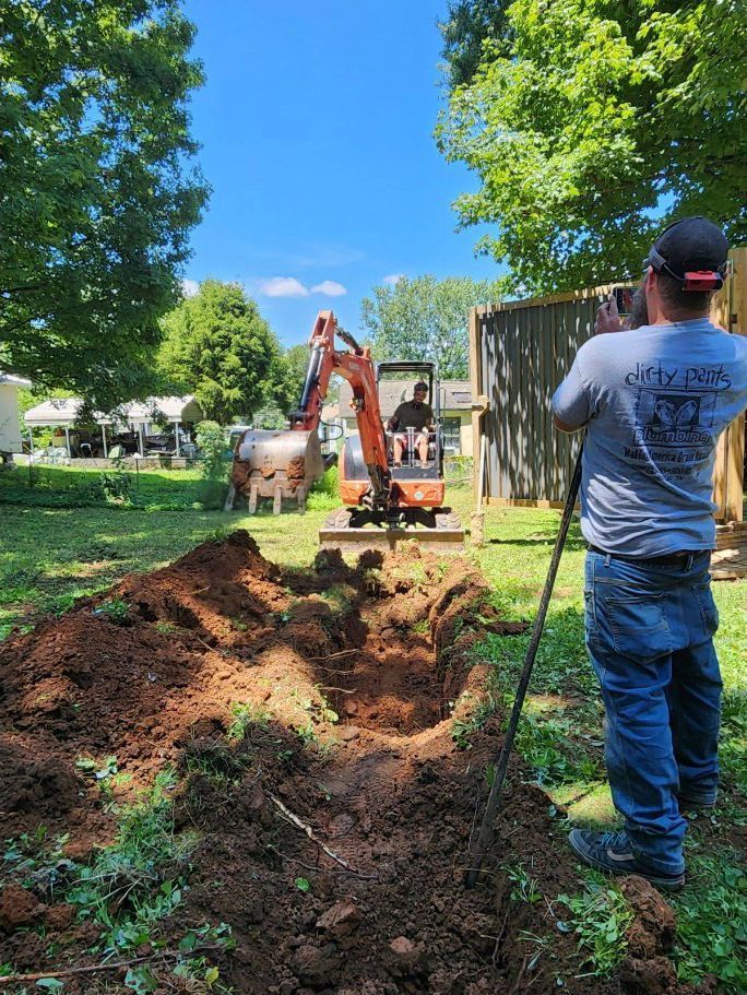 Worker operates a mini excavator digging a trench in a grassy yard beside a shed.