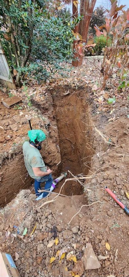 Worker in green helmet digging a deep trench in an outdoor dirt area with trees and shrubs.