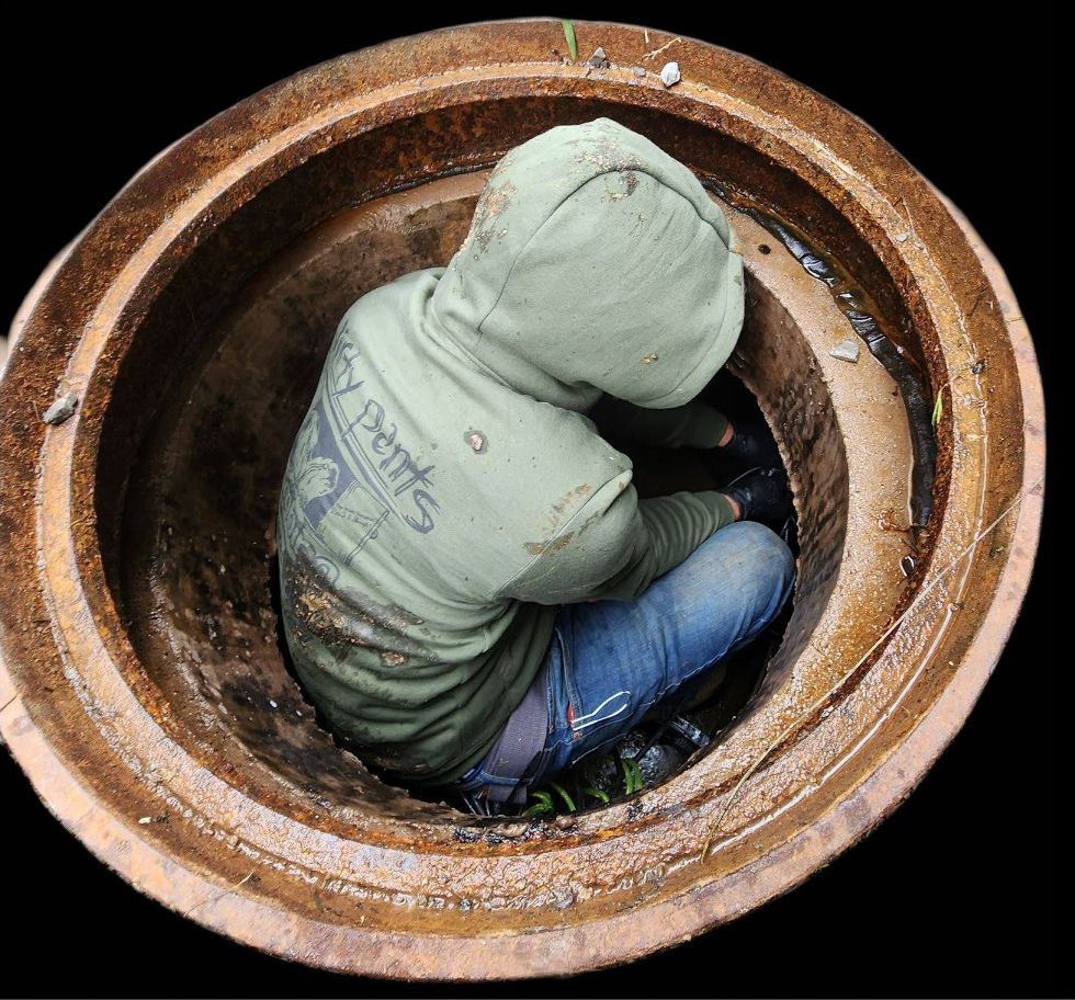 Person crouched inside a large rusty circular pipe, wearing a light green hoodie and blue jeans.