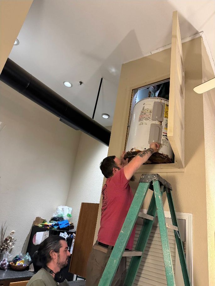 Person on a green ladder working inside a wall opening while another person looks on nearby.