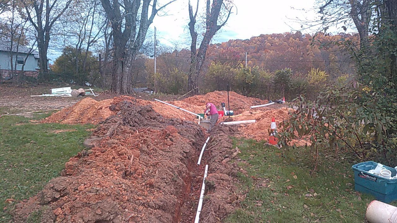 Dirt trench in a grassy yard with a white pipe and a person in pink near the far end