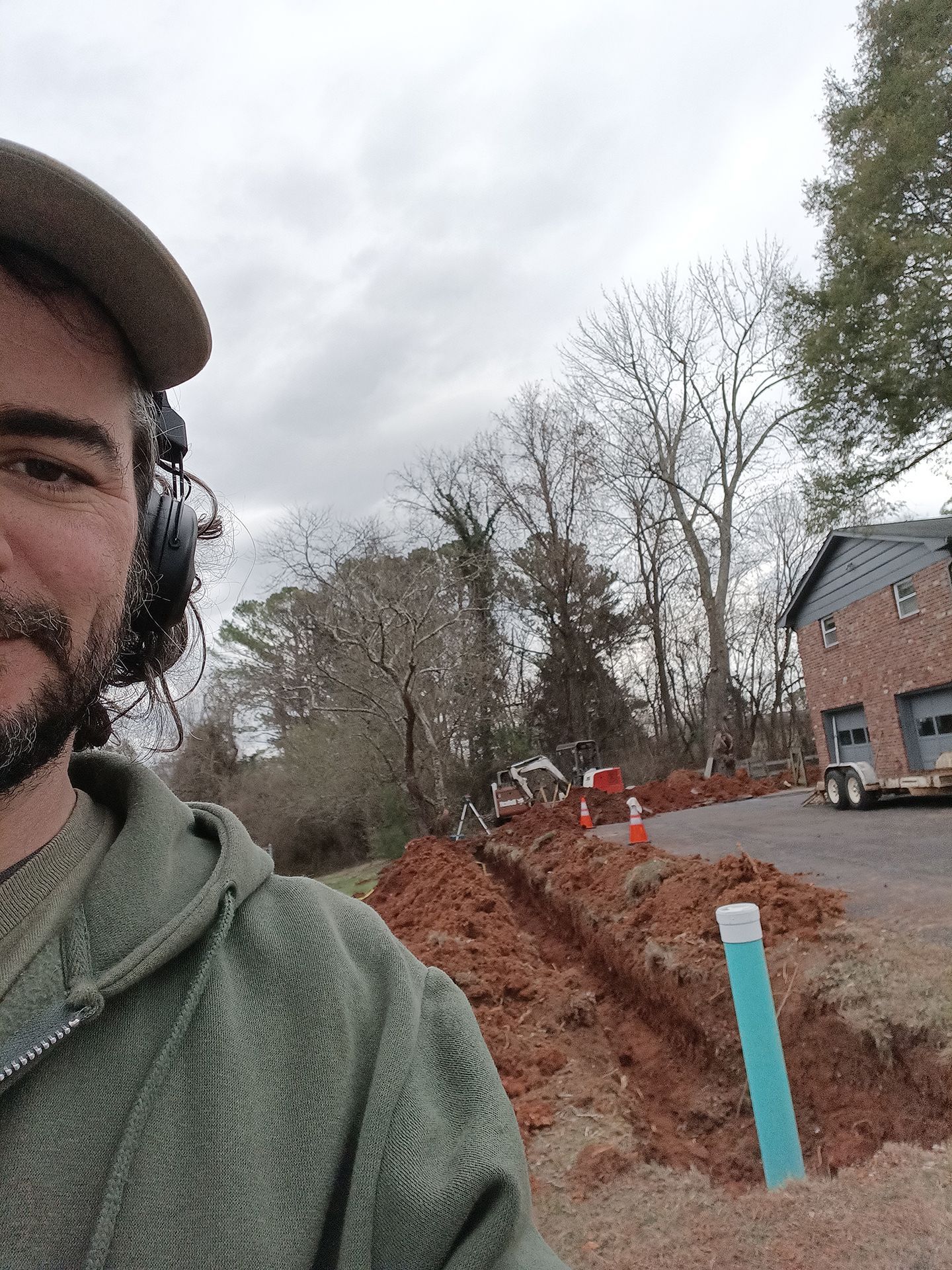 Selfie of a man on a muddy roadside with a trench, blue marker post, and houses under a gray sky