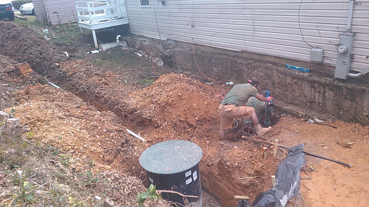 Workers clearing dirt beside a house, with a trench, mulch pile, and construction equipment.