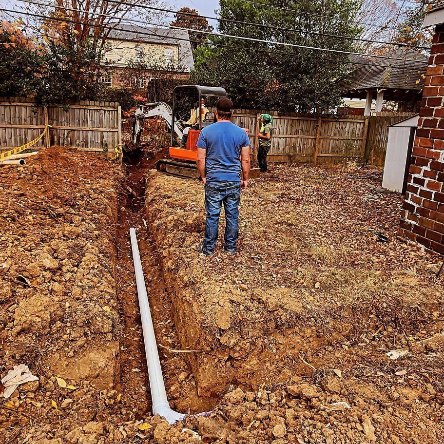 Person in blue shirt standing in a backyard beside a trench and orange excavator, with a white pipe in the foreground.