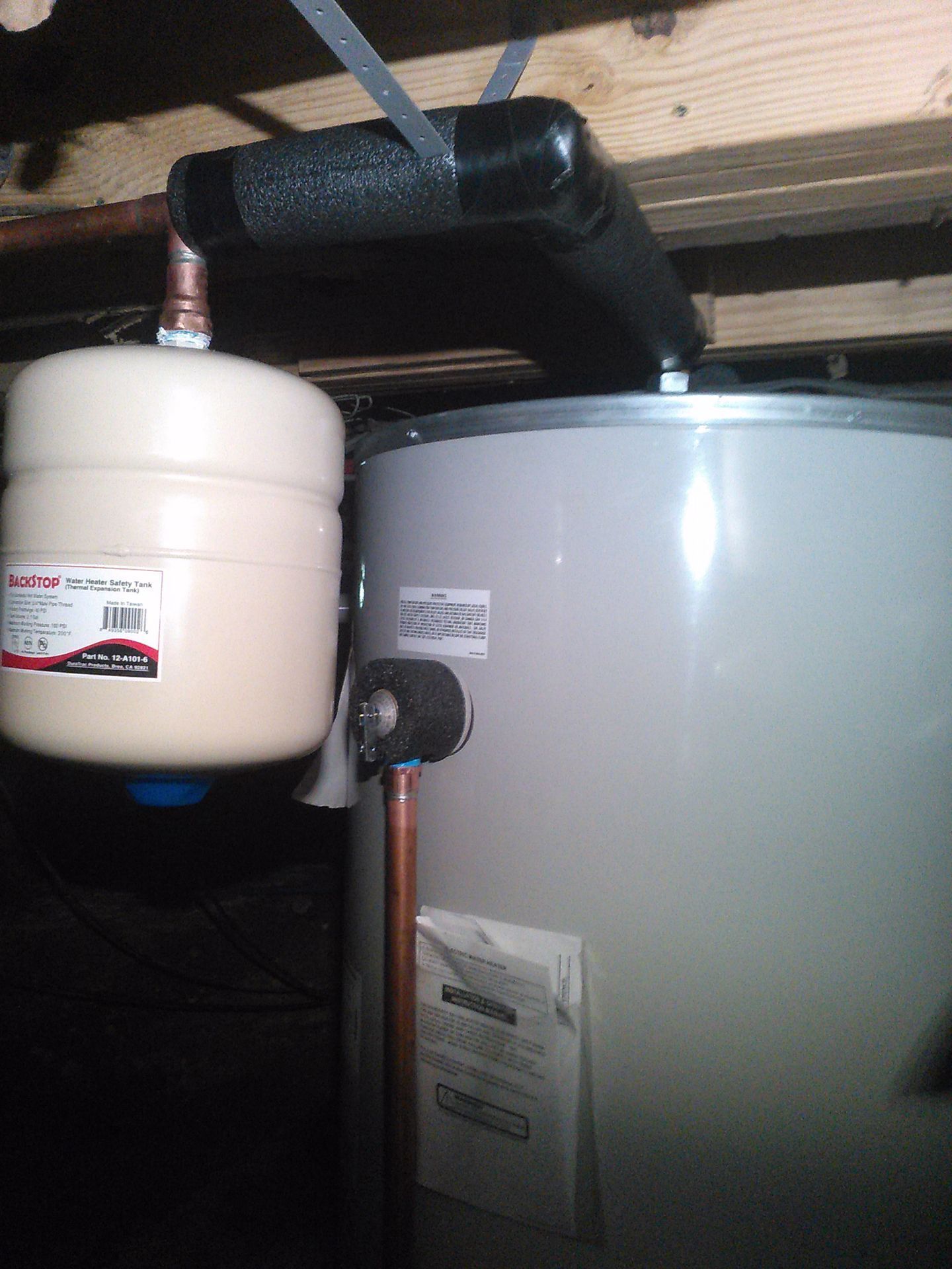 White expansion tank and gray water heater with black insulated pipes in a basement.
