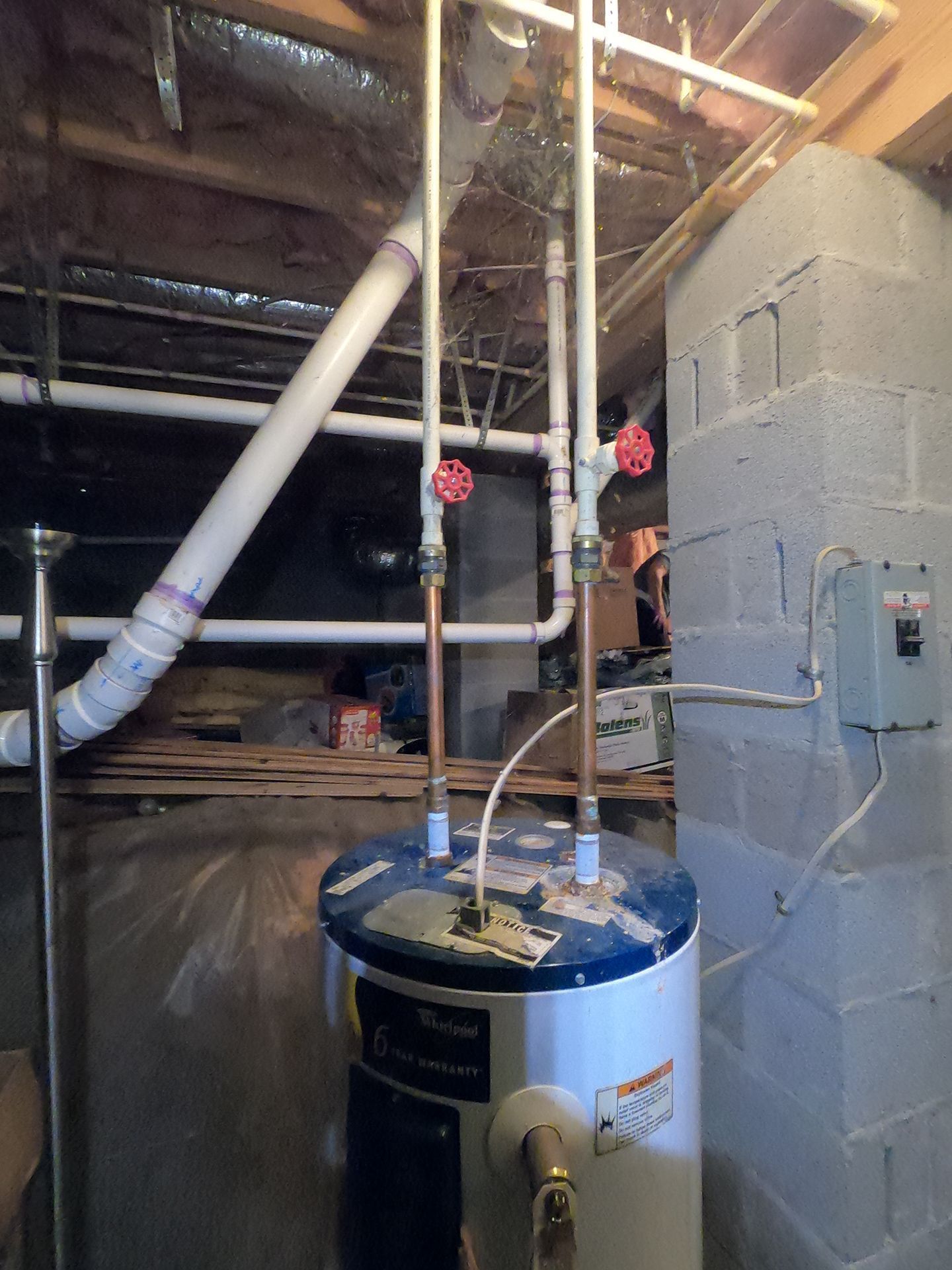 Water heater and exposed pipes in a basement, with a worker standing on a ladder near ductwork.