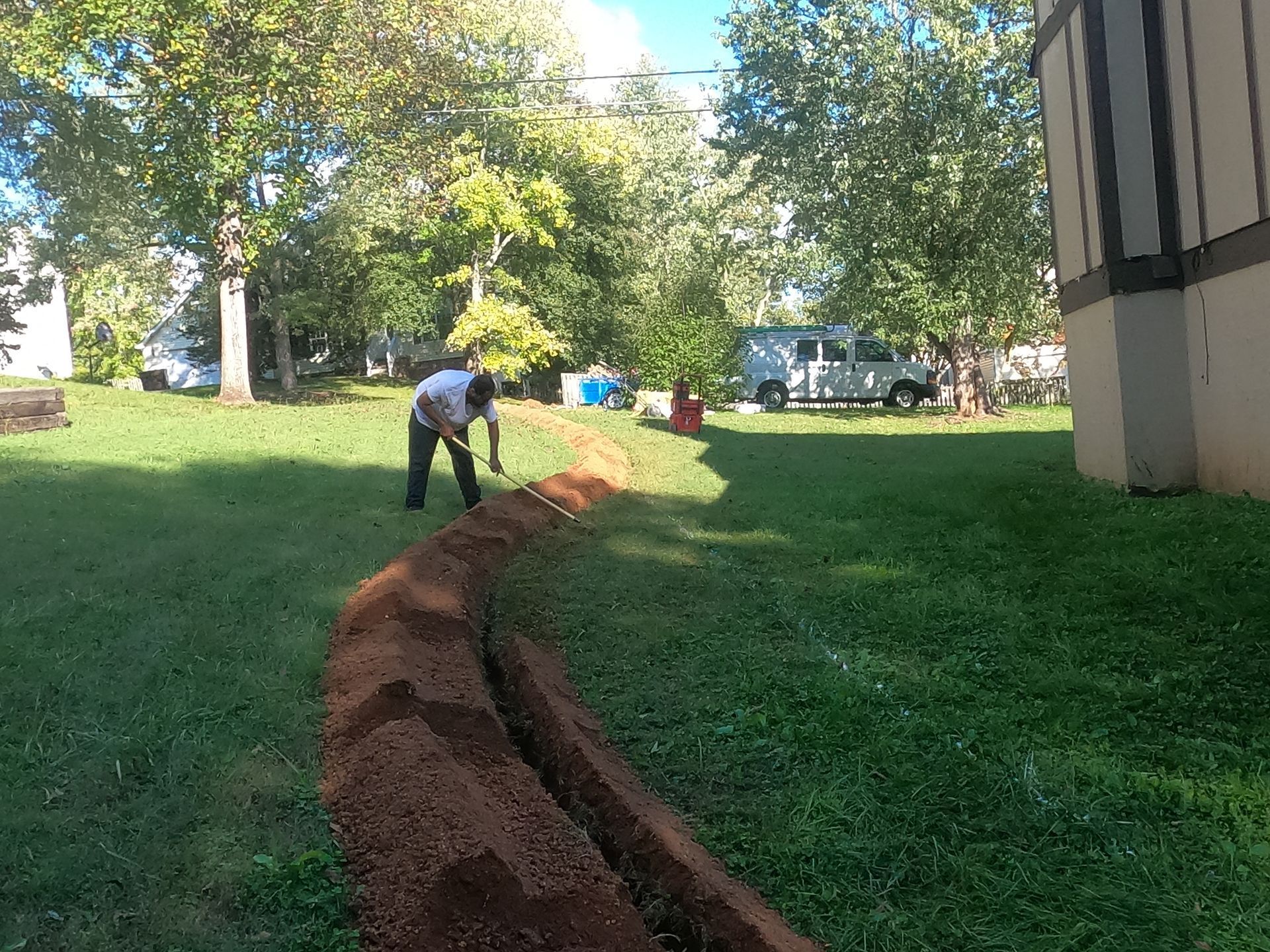 Worker digging a narrow trench through a grassy yard beside a building on a sunny day