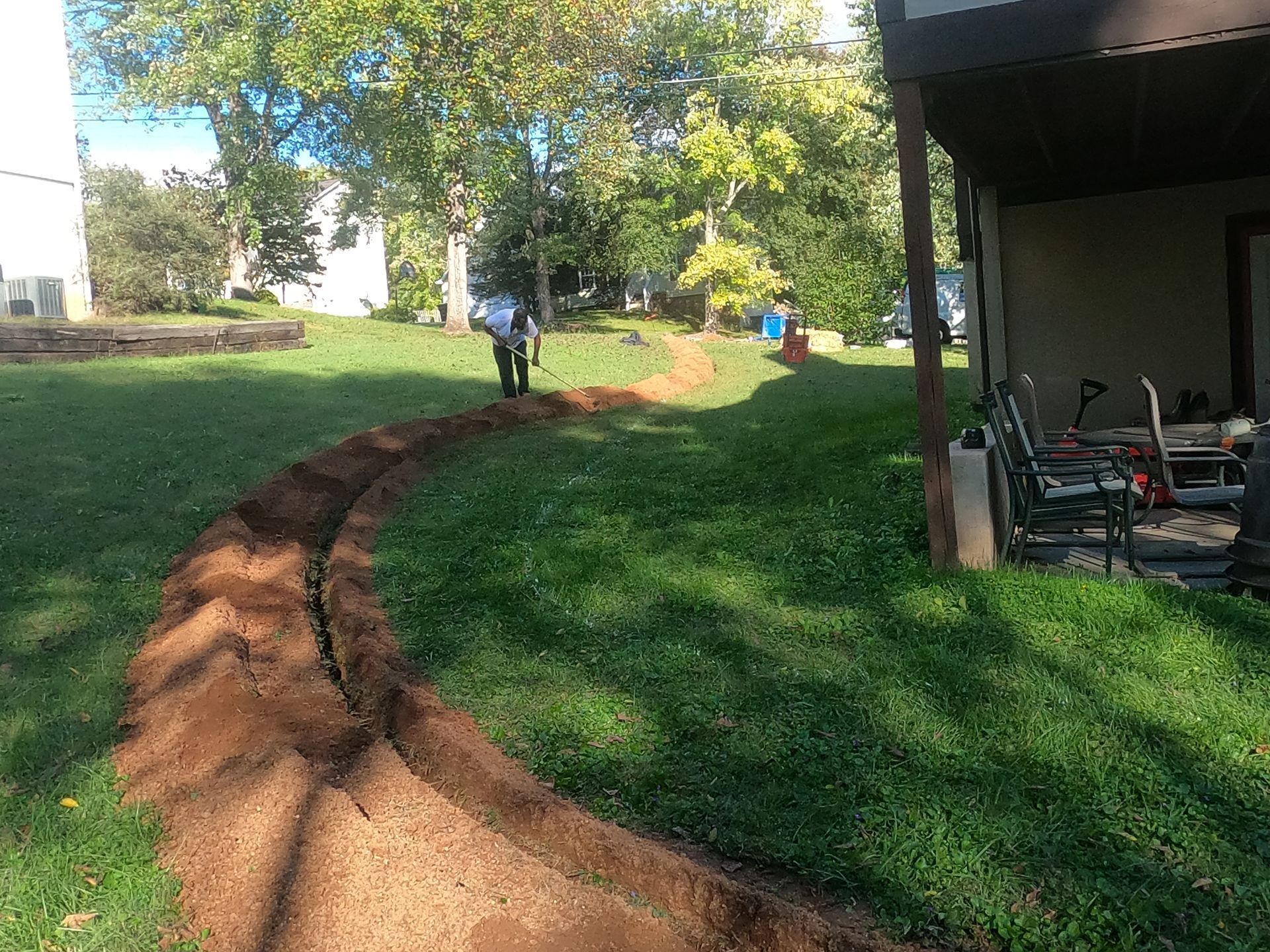 Dirt path through grassy yard with trees and a person in the distance beside a house and porch.