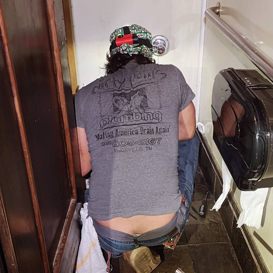 Person crouching in a bathroom beside a toilet and wall-mounted dispenser, wearing a gray shirt and cap.
