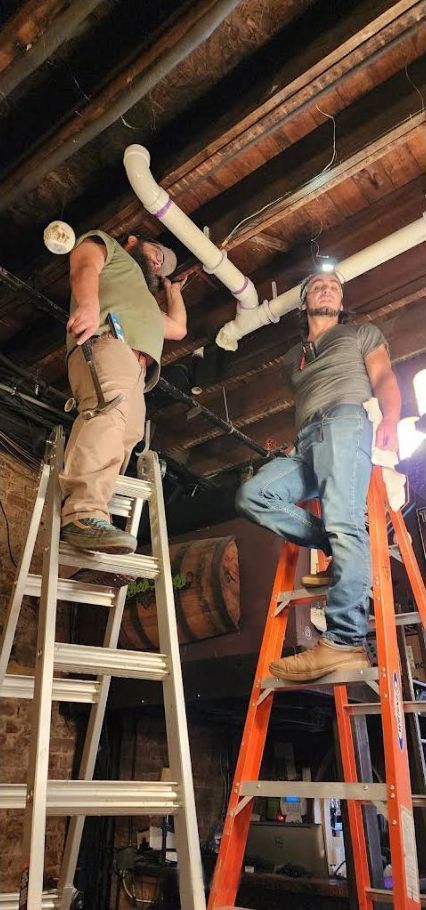 Two workers on ladders installing ductwork in an unfinished ceiling room
