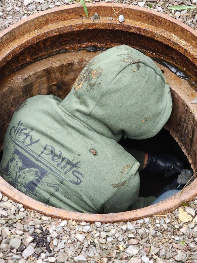 Two workers in green hoodies crouch inside an open manhole on gravel ground.