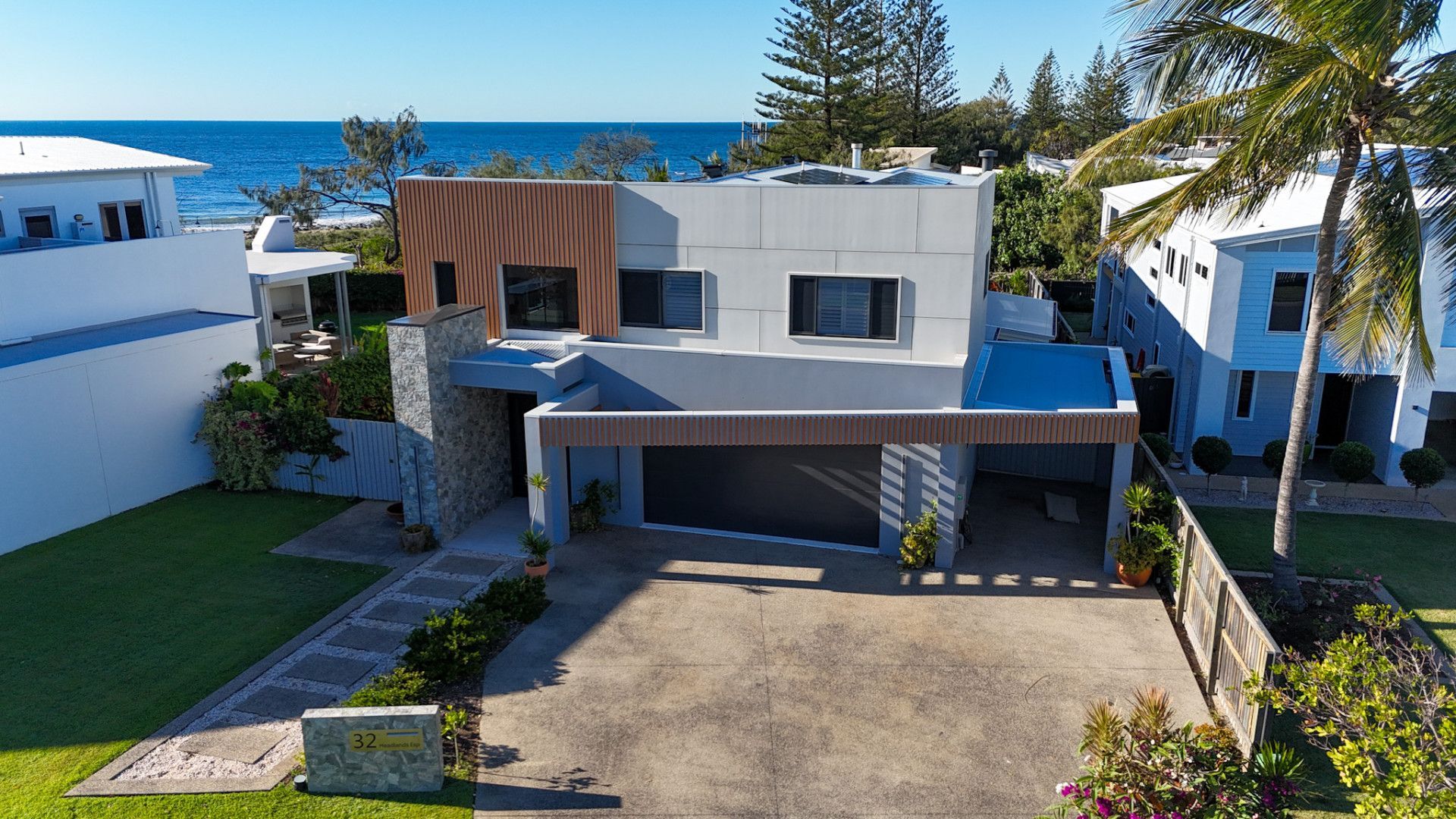 Modern beachfront house with a driveway, ocean visible behind it, and palm trees nearby