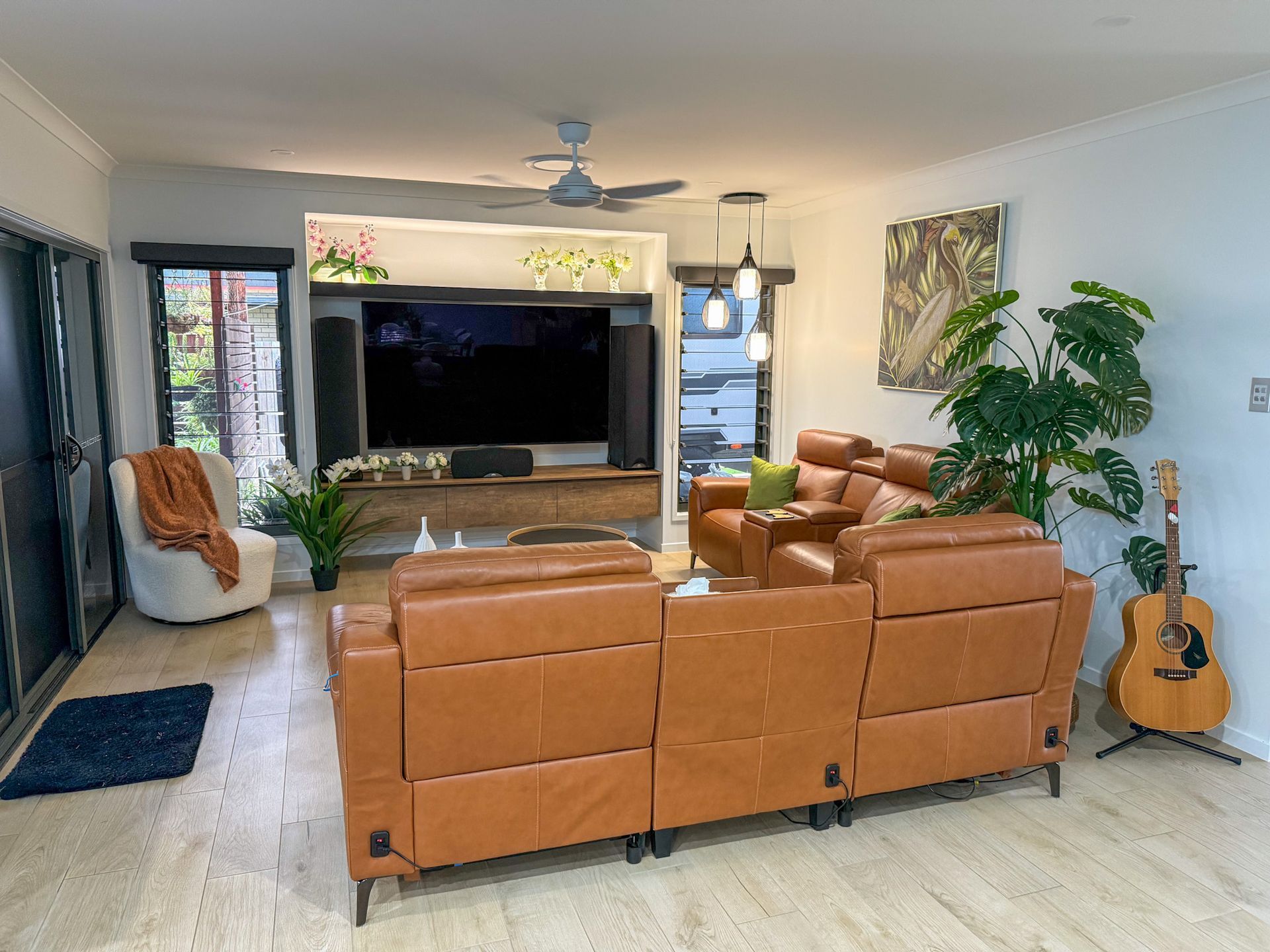 Bright living room with tan sectional, large TV, guitar, plants, and open doors to the outside