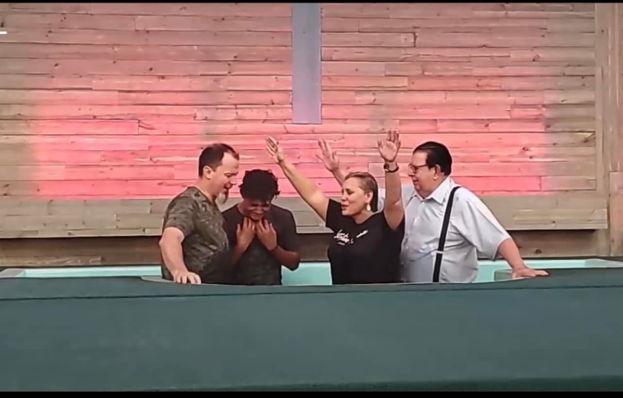 People in a baptismal pool, one raising arms, others smiling. Red light on wood background.