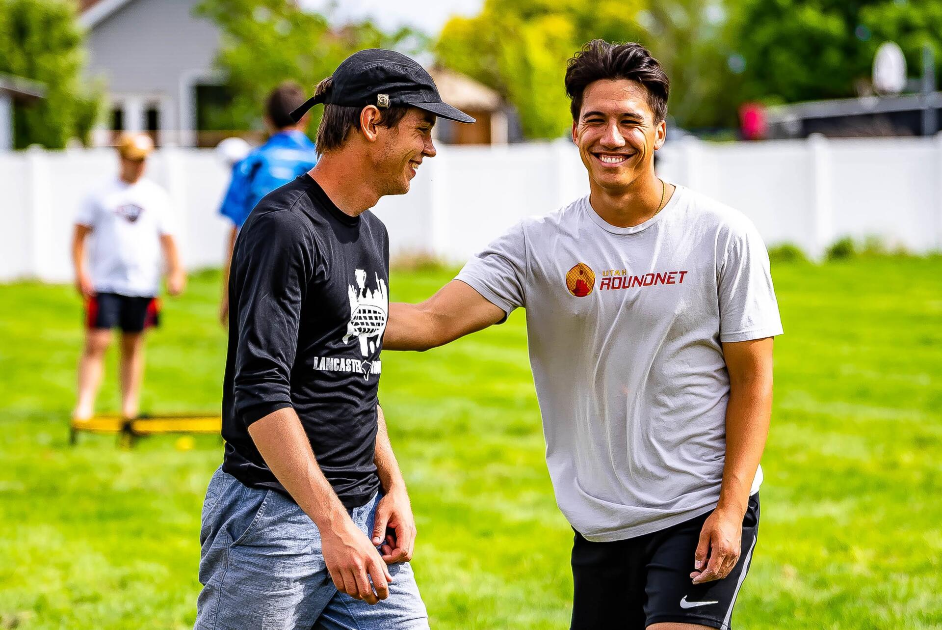 Two men smiling on a grassy field. One has his hand on the other's shoulder. Others in background.
