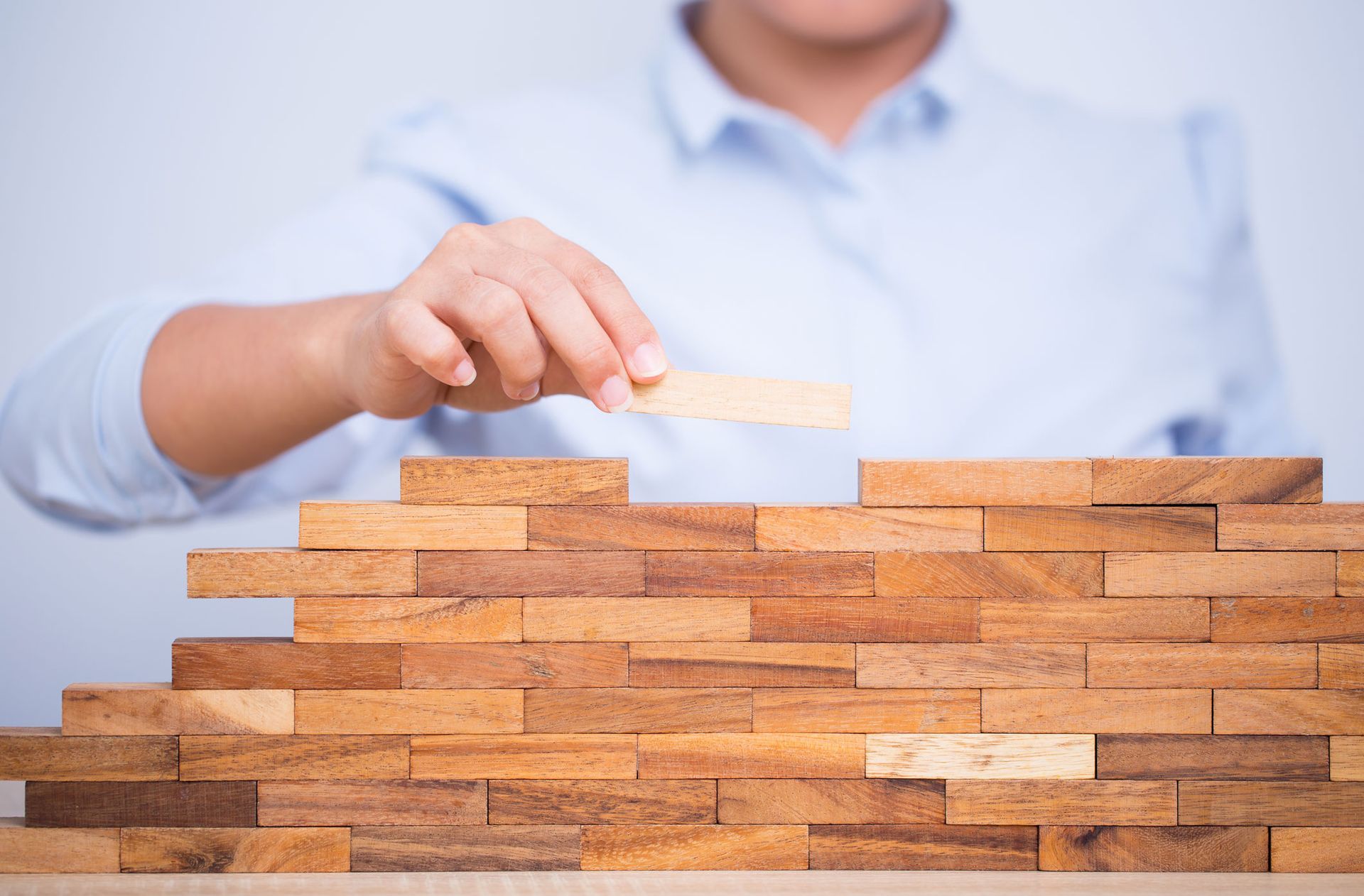 Person placing a wooden block on top of a partially built wall, symbolizing construction. Person placing a wooden block on top of a partially built wall, symbolizing construction.