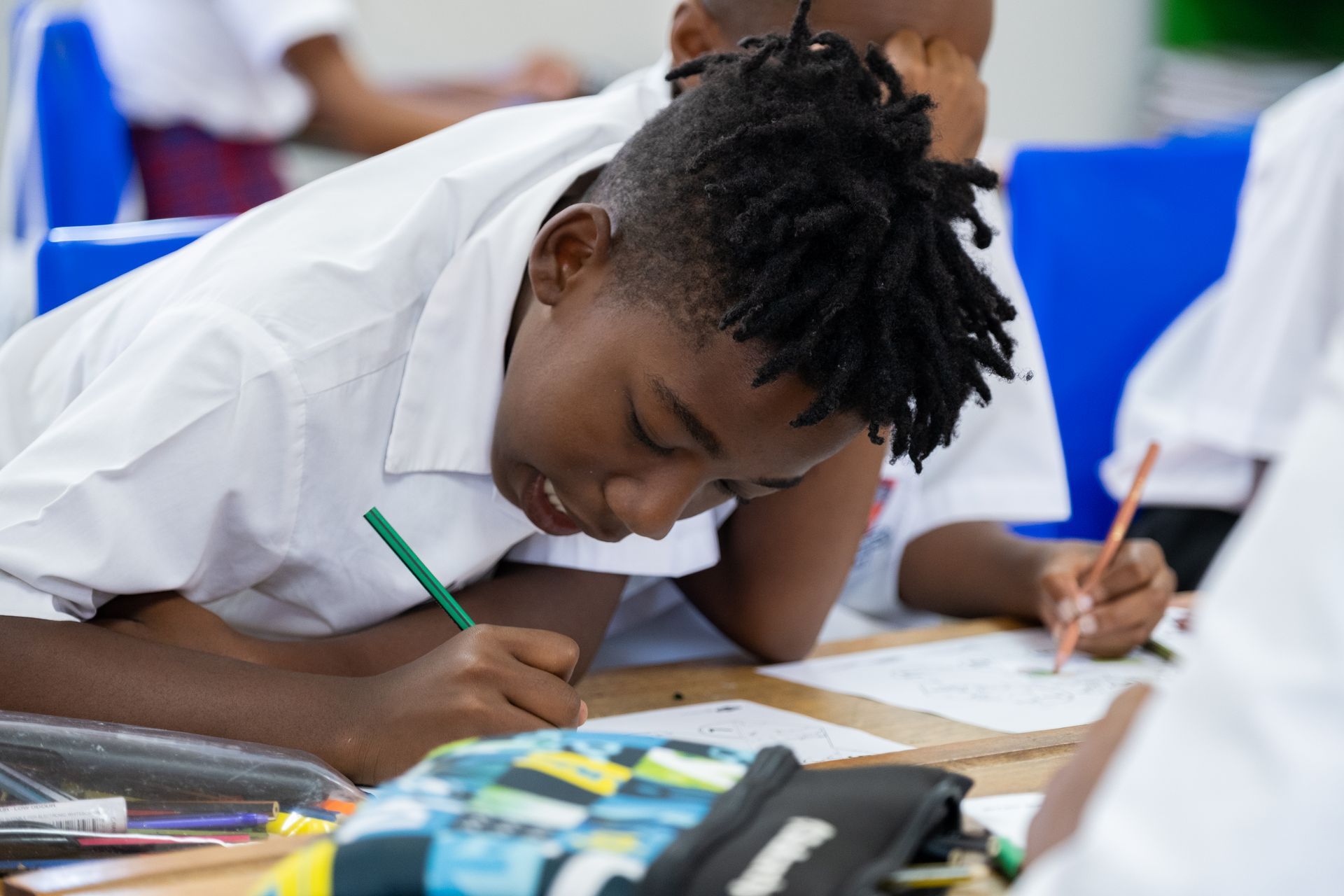 A young boy is sitting at a desk in a classroom writing on a piece of paper.