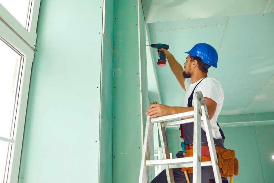 Worker on a ladder drilling a teal wall indoors, wearing a blue hard hat and white shirt.