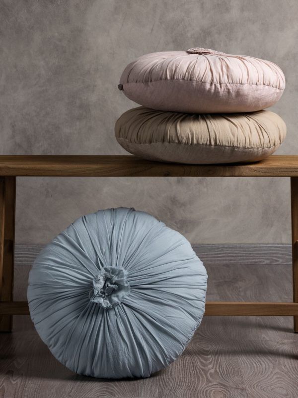 Tre cuscini rotondi in tessuto plissettato: uno rosa chiaro e uno beige, uno sopra l'altro, e uno blu su una panca.