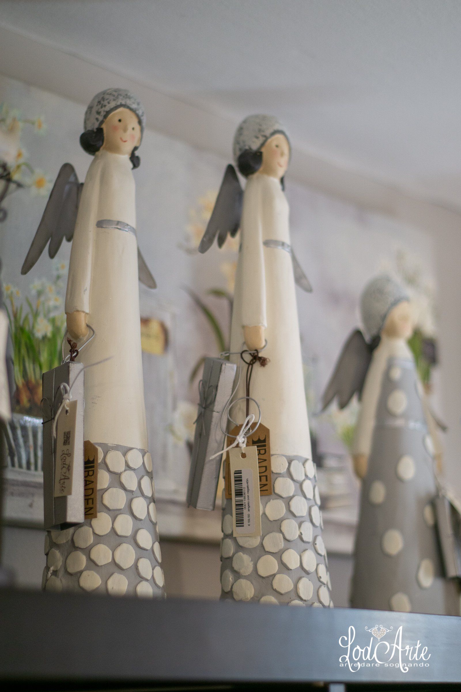 Tre statuette di angeli in ceramica su una mensola, con abiti bianchi e grigi, ali ed etichette.