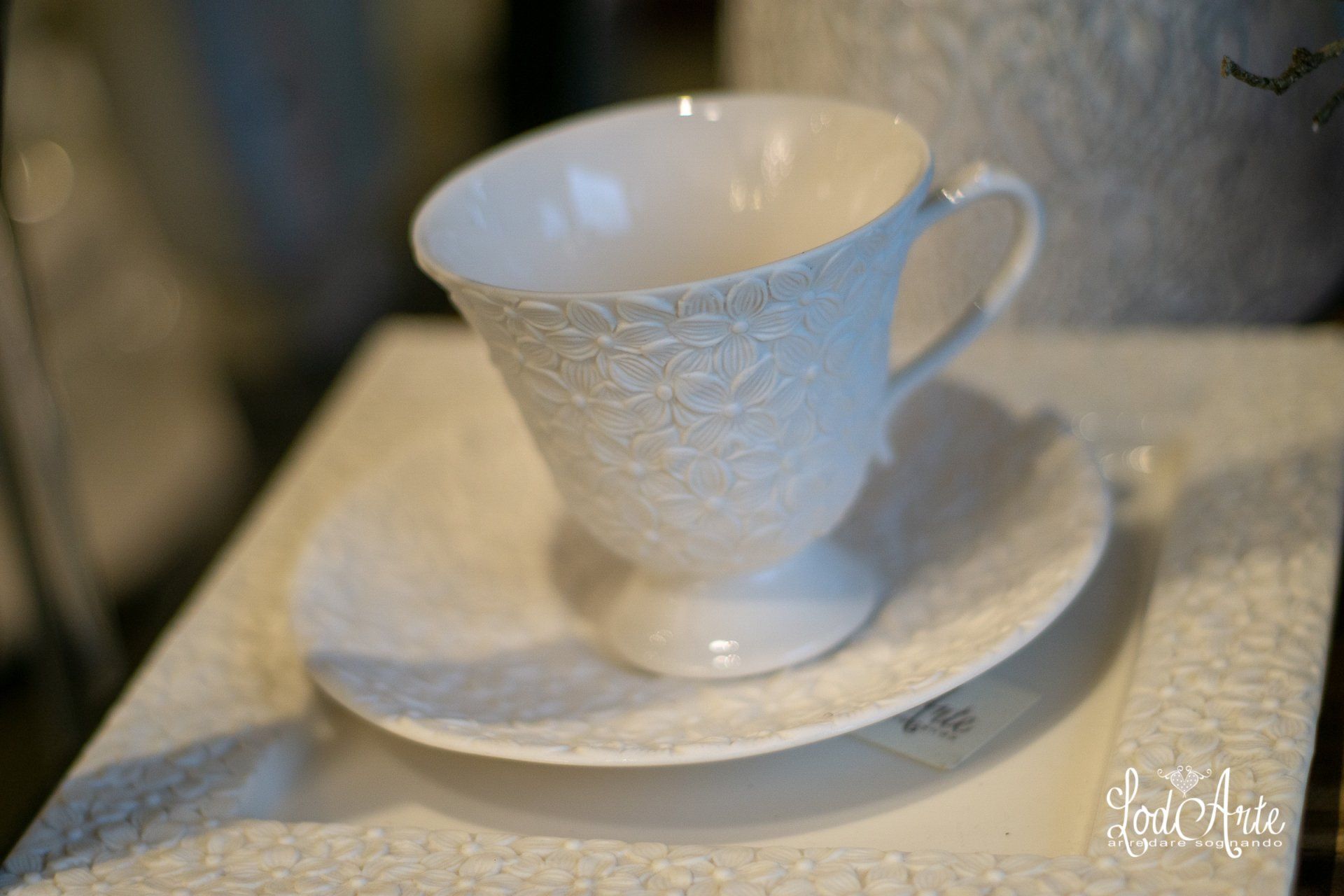 Tazza da tè bianca con piattino con intricato motivo in rilievo su un piatto quadrato testurizzato.