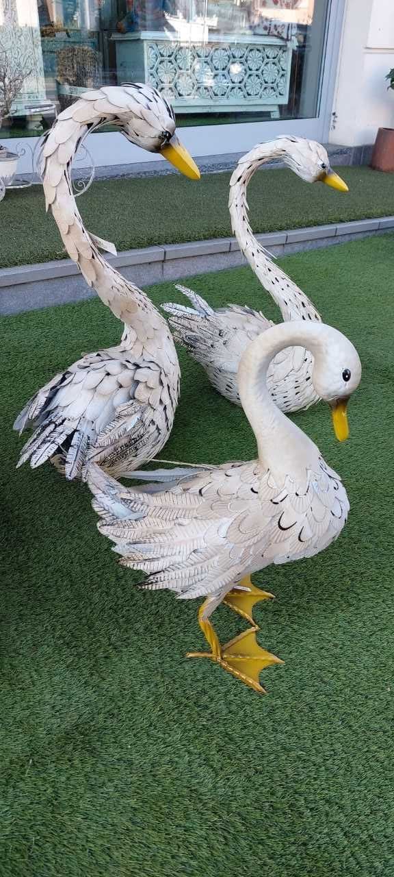 Tre statue di cigni bianchi con macchie nere e becco e zampe gialli su erba verde.