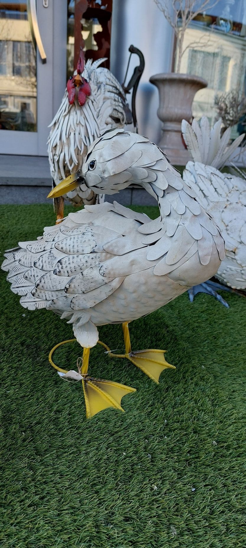 Scultura di un'anatra in metallo bianco con zampe gialle, con scultura di un gallo sullo sfondo su erba artificiale verde.