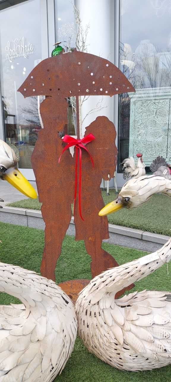 Scultura in metallo raffigurante due figure sotto un ombrello, con un fiocco. Cigni in primo piano, adagiati sull'erba.