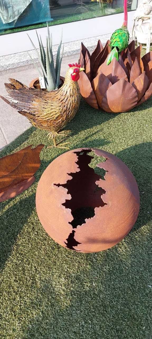 Una fioriera per galline con una pianta all'interno, un vaso di fiori in metallo e una sfera crepata su erba verde.