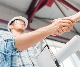 Construction worker in a hard hat and glasses shaking hands with someone; indoor construction site.