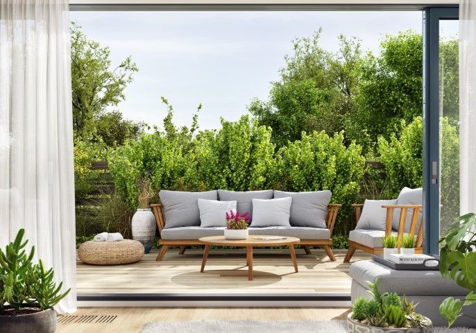 Modern patio with gray sofa, white cushions, and lush green garden view through open sliding doors