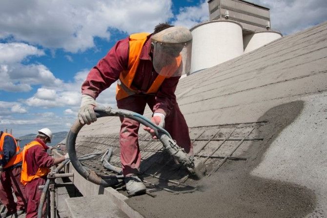 Workers on a rooftop applying gray roofing material with hoses under a partly cloudy sky