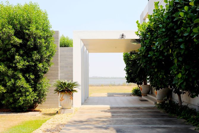 Sunlit white courtyard entrance framed by green hedges and potted plants, leading to open fields beyond