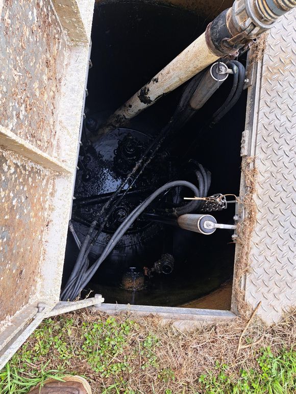 Open manhole with dark water, hoses, and a pump inside near a grassy edge.