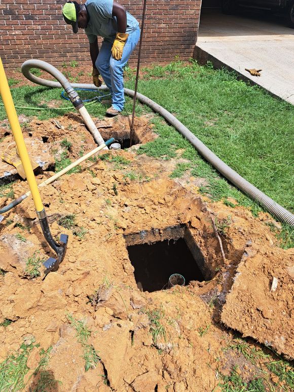 Worker standing beside an open utility trench with hoses and muddy soil near a brick wall.