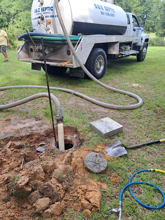 White septic truck pumping into an open ground hole beside a grassy yard and hoses.