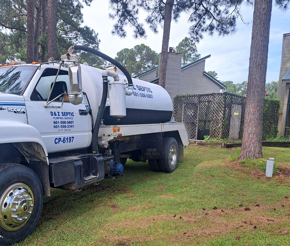 White utility truck parked on grass beside a stone building and tall pine trees.