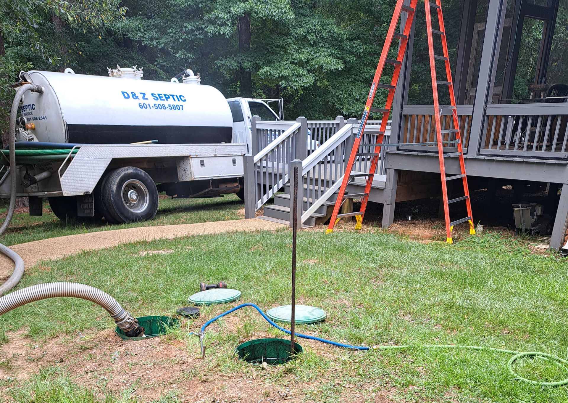 Yard scene with a water tanker, ladder, trailer, and hoses around a tree stump