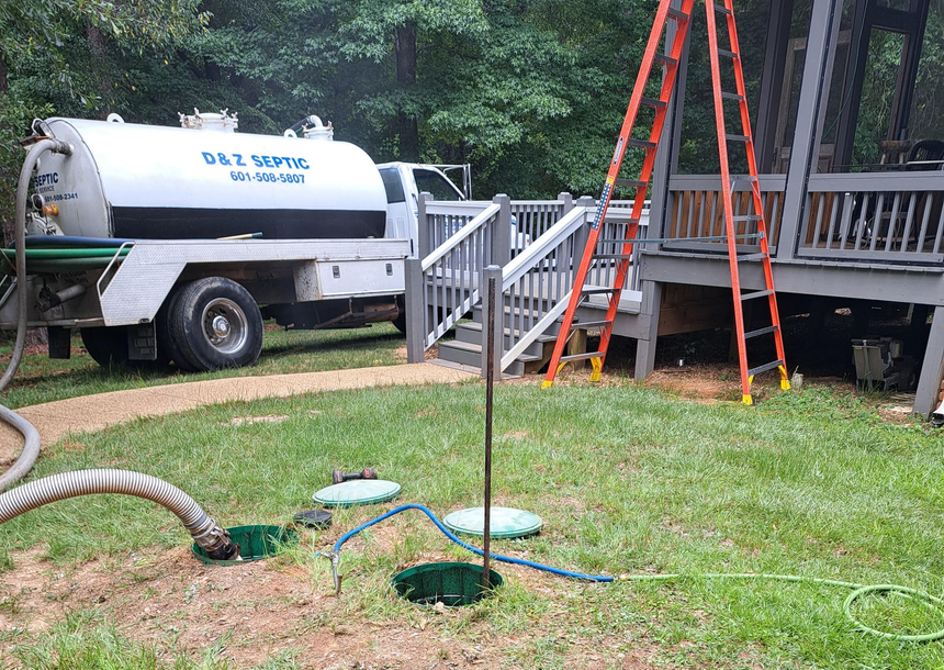 Water truck beside a house deck with orange ladder, hoses on grass, and a tank opening in the yard