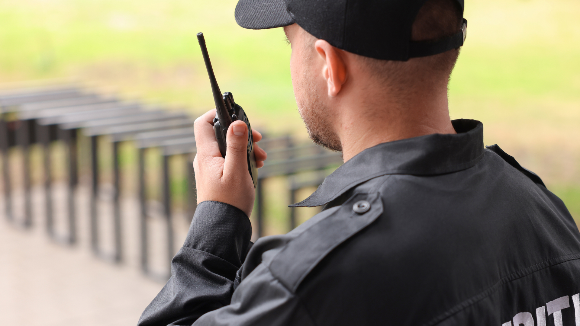 Security guard in black uniform using a walkie-talkie outdoors near a fence.