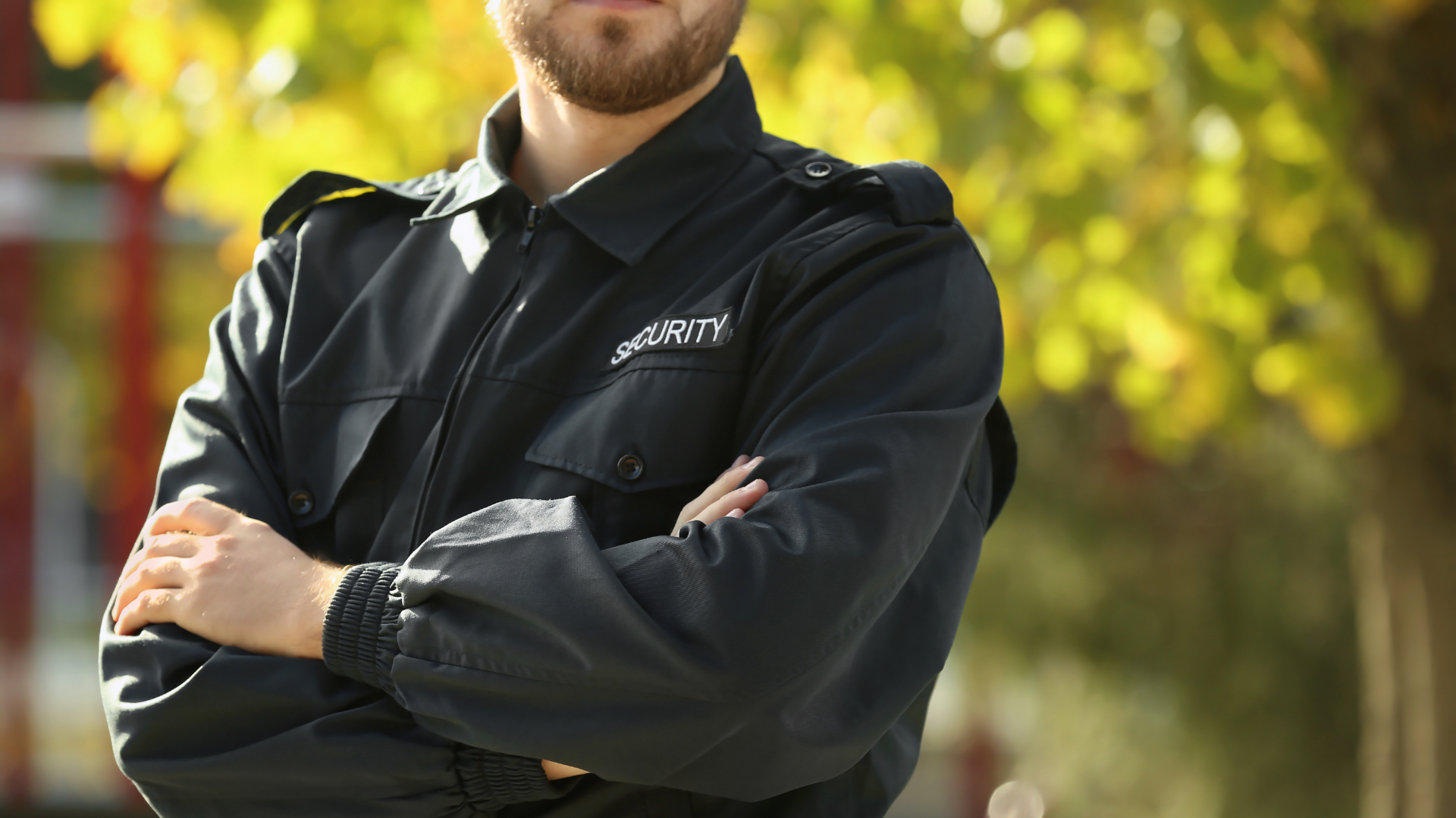 Person in a black jacket standing with arms crossed outdoors against a blurred yellow-green background