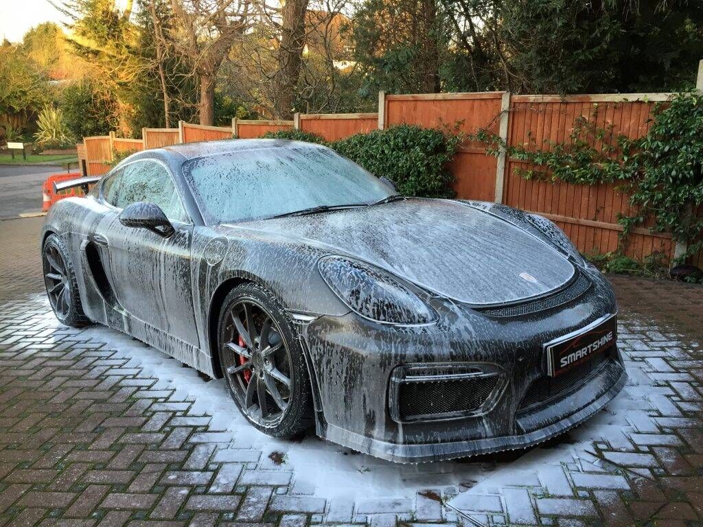 Black sports car covered in foam on a driveway, being washed outdoors ליד wooden fence